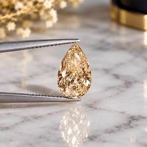 May include: A pear-shaped champagne-coloured gemstone held by silver-toned metal tweezers. The faceted stone exhibits a warm, golden-brown hue with intricate light-reflecting surfaces, showcasing a brilliant cut that creates a sharp, pointed top.