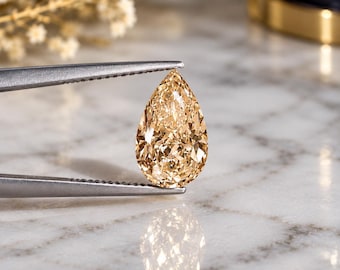 May include: A pear-shaped champagne-colored gemstone held by silver-toned metal tweezers. The faceted stone exhibits a warm, golden-brown hue with intricate light-reflecting surfaces, showcasing a brilliant cut that creates a sharp, pointed top.