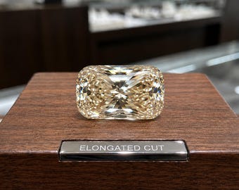 May include: A faceted, elongated cushion-cut gemstone with a light champagne hue, resting on a dark brown wooden display block. A small, rectangular silver-toned metal plaque is embedded in the wood, featuring the engraved text ELONGATED CUT.