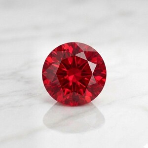 May include: A round, brilliant-cut gemstone with a deep, vibrant red color. The stone features numerous triangular and kite-shaped facets that create a complex, symmetrical pattern of light reflection and refraction across its circular surface.