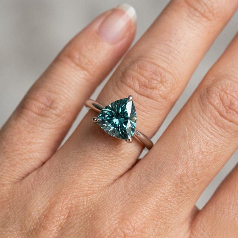 May include: A silver-toned ring featuring a large, faceted, trillion-cut teal gemstone held in place by a three-prong setting. The band is smooth and polished, showcasing a clean, minimalist design that highlights the triangular stone.