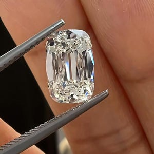 Peut inclure: Une pierre précieuse incolore et transparente, taillée en coussin avec une surface à facettes qui reflète la lumière, tenue par une pince en métal gris. La pierre présente une forme rectangulaire aux coins arrondis et une structure interne brillante.
