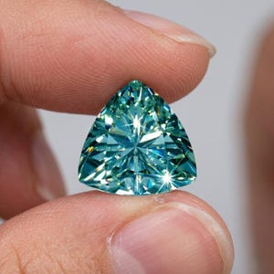 May include: A trillion-cut gemstone with a vibrant teal-blue color and a highly faceted surface that reflects light with intense brilliance. The triangular stone features sharp, clean edges and a symmetrical arrangement of facets throughout its body.