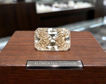 May include: A faceted, champagne-colored gemstone with an elongated cushion cut, resting on a dark brown wooden display block. A small, rectangular silver-toned metal plaque is embedded in the wood, featuring the engraved text ELONGATED CUSHION.