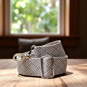 May include: A light grey and white patterned belt with a silver-coloured buckle and clasp. The belt is made of a woven fabric and is lying on a wooden surface. The background includes a window and a pillow.