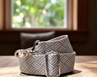 May include: A light gray and white patterned belt with a silver-colored buckle and clasp. The belt is made of a woven fabric and is lying on a wooden surface. The background includes a window and a pillow.
