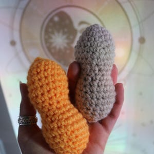 May include: Two handmade crochet peanut shapes. One is bright orange, the other is gray. The peanuts are held in a hand, with a silver ring on the finger. The background is a soft, blurred image of a celestial design.