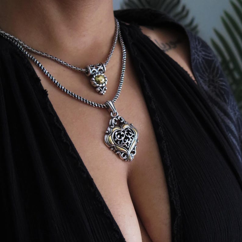 Op de afbeelding: Twee zilveren kettingen met sierlijke hangers. De onderste hanger is hartvormig met gouden accenten en ingewikkelde filigraan. De bovenste hanger is kleiner, met een gouden middenstuk. Beide kettingen hebben een getextureerde ketting.