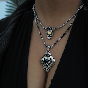 Op de afbeelding: Twee zilveren kettingen met sierlijke hangers. De bovenste ketting heeft een kleine, ronde hanger met gouden accenten. De onderste ketting heeft een grotere, ruitvormige hanger met gouden filigraan en een zwarte achtergrond.