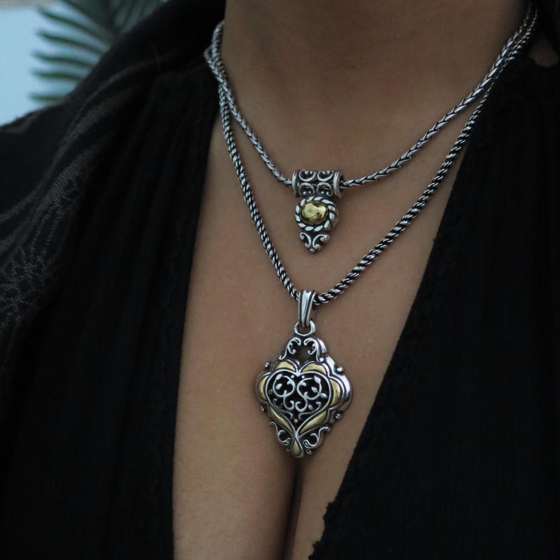 Op de afbeelding: Twee zilveren kettingen met sierlijke hangers. De bovenste ketting heeft een kleine, ronde hanger met gouden accenten. De onderste ketting heeft een grotere, ruitvormige hanger met gouden filigraan en een zwarte achtergrond.