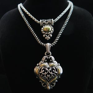 Op de afbeelding: Twee zilveren kettingen met ingewikkelde ontwerpen. De bovenste ketting heeft een hanger met een goudkleurig midden en sierlijke zilveren details. De onderste ketting heeft een grotere hanger met een goud-zilveren filigraan ontwerp. Beide kettingen worden tegen een zwarte achtergrond getoond.