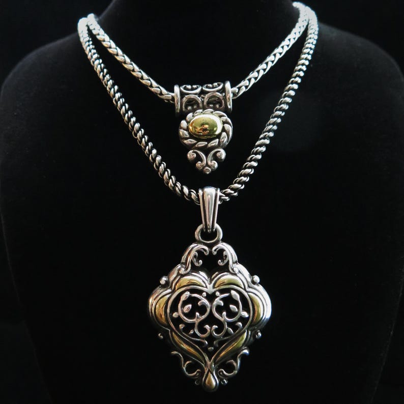 Op de afbeelding: Twee zilveren kettingen met ingewikkelde ontwerpen. De bovenste ketting heeft een hanger met een goudkleurig midden en sierlijke zilveren details. De onderste ketting heeft een grotere hanger met een goud-zilveren filigraan ontwerp. Beide kettingen worden tegen een zwarte achtergrond getoond.