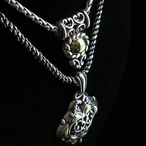 Op de afbeelding: Twee zilveren kettingen met sierlijke hangers. De bovenste ketting heeft een cilindrisch accent met een goudkleurig midden. De onderste ketting heeft een grotere, gedetailleerde hanger met gouden accenten. De achtergrond is zwart.