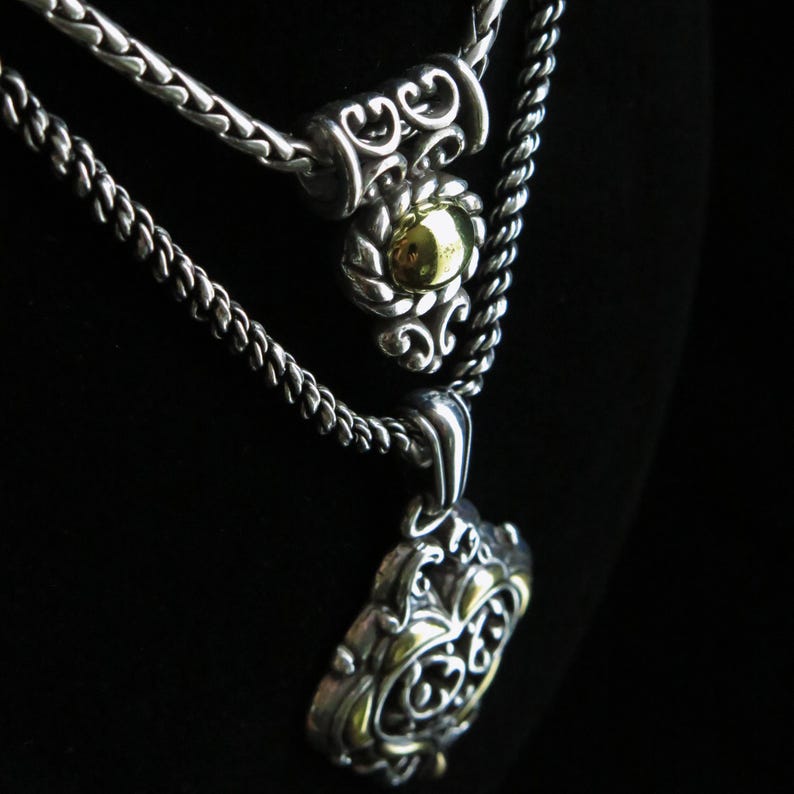 Op de afbeelding: Twee zilveren kettingen met sierlijke hangers. De bovenste ketting heeft een cilindrisch accent met een goudkleurig midden. De onderste ketting heeft een grotere, gedetailleerde hanger met gouden accenten. De achtergrond is zwart.