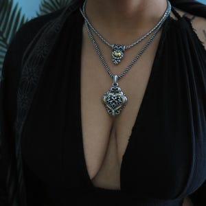 Op de afbeelding: Twee zilveren kettingen met sierlijke hangers. De bovenste hanger is klein en rond met een gouden centrum. De onderste hanger is een groter, sierlijk ontwerp met zilveren en gouden accenten. De kettingen zijn gelaagd en gedragen op een zwarte top.