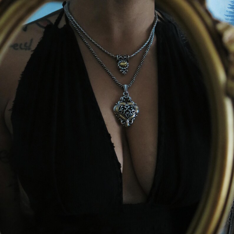 Op de afbeelding: Twee zilveren kettingen met sierlijke hangers. De bovenste ketting heeft een kleine, ronde hanger met een goudkleurig midden. De onderste ketting heeft een grotere, hartvormige hanger met ingewikkelde filigraan details. De kettingen worden tegen een zwarte achtergrond getoond.