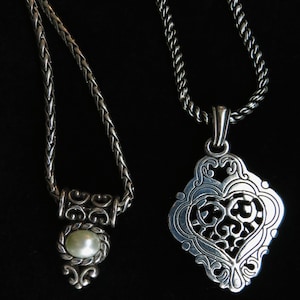 Op de afbeelding: Twee zilveren kettingen met ingewikkelde ontwerpen. E&eacute;n heeft een gevlochten ketting met een hanger met een parel. De andere heeft een decoratieve hanger met een hartvormig filigraanontwerp, hangend aan een gedraaide ketting.