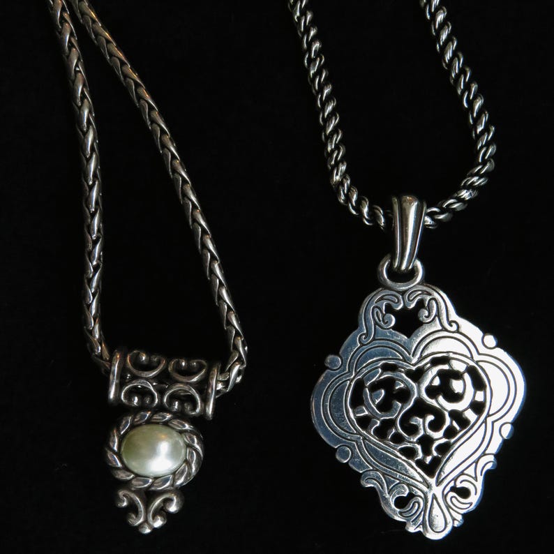 Op de afbeelding: Twee zilveren kettingen met ingewikkelde ontwerpen. E&eacute;n heeft een gevlochten ketting met een hanger met een parel. De andere heeft een decoratieve hanger met een hartvormig filigraanontwerp, hangend aan een gedraaide ketting.