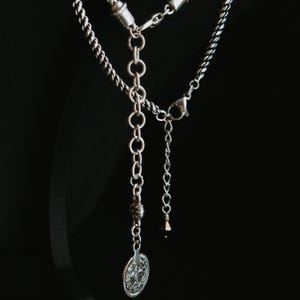 Op de afbeelding: Zilverkleurige ketting met een gedetailleerde hanger en verschillende kettingstijlen. De ketting heeft een ronde hanger met een reli&euml;f ontwerp, een kralen accent en een karabijnsluiting. De achtergrond is zwart.