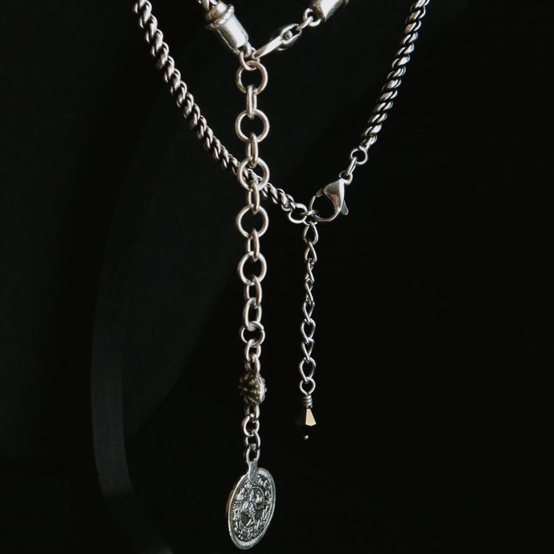 Op de afbeelding: Zilverkleurige ketting met een gedetailleerde hanger en verschillende kettingstijlen. De ketting heeft een ronde hanger met een reli&euml;f ontwerp, een kralen accent en een karabijnsluiting. De achtergrond is zwart.