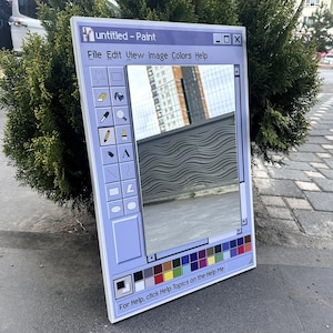 Op de afbeelding: Een rechthoekige spiegel omlijst om te lijken op het klassieke "Untitled - Paint" toepassingsvenster. Het lichtblauwe frame heeft de titelbalk van het programma, menu-opties en een kleurenpalet. De spiegel reflecteert de omgeving.