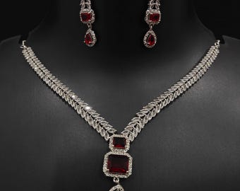 Puede incluir: Un conjunto de collar y pendientes plateados con detalles de piedras preciosas rojas. El collar presenta un colgante con una piedra rectangular grande y una piedra en forma de lágrima. Los pendientes tienen un diseño similar.