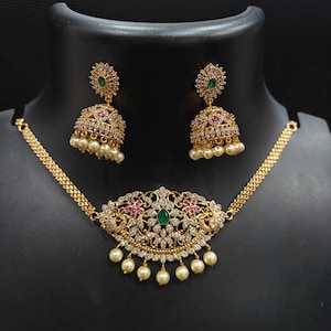 Puede incluir: Un conjunto de joyas dorado que incluye un collar y pendientes a juego. El collar presenta un diseño floral central con una piedra verde, rodeada de piedras claras y rojas. Los pendientes y el collar están adornados con pequeñas perlas colgantes.