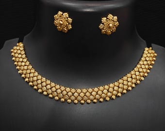 May include: A gold-colored jewelry set featuring a necklace and matching earrings. The necklace has a beaded design with a curved shape. The earrings are flower-shaped. The jewelry is displayed on a black mannequin.