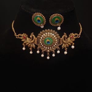 Op de afbeelding: Een goudkleurige ketting- en oorbellenset. De ketting heeft een centraal pauwenontwerp met een groen en blauw oog, omringd door witte edelstenen. De oorbellen spiegelen het pauwenoogontwerp. Kleine witte parels hangen aan de ketting.