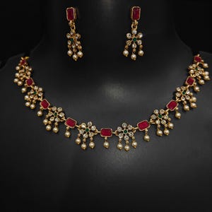 Puede incluir: Un conjunto de joyas dorado con un collar y pendientes a juego. El collar y los pendientes están adornados con piedras rectangulares rojas, pequeñas perlas blancas y racimos de piedras transparentes y verdes. El conjunto se exhibe sobre un maniquí negro.