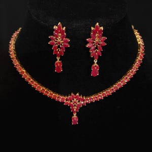 May include: A gold-toned necklace and earring set. The necklace and earrings feature ruby-red gemstones. The necklace has a floral pendant and the earrings are a matching floral design. The jewelry is displayed on a black background.
