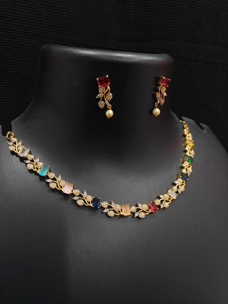 Op de afbeelding: Een goudkleurige ketting- en oorbellenset. De ketting heeft kleurrijke rechthoekige edelstenen en bladaccenten. De oorbellen hebben een rode edelsteen en een bungelende parel. De sieraden worden op een zwarte mannequin getoond.