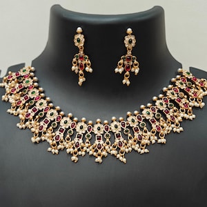 Puede incluir: Juego de collar y pendientes dorados. El collar y los pendientes presentan piedras preciosas rojas y verdes, pequeñas perlas blancas y un diseño floral. Las joyas se exhiben en un maniquí negro.