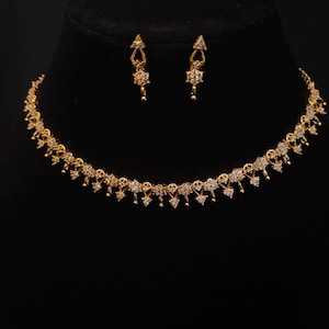 Puede incluir: Un conjunto de collar y pendientes de color dorado. El collar y los pendientes presentan un diseño delicado con pequeños acentos brillantes. Las joyas se exhiben sobre un fondo negro, resaltando el color dorado y los intrincados detalles.