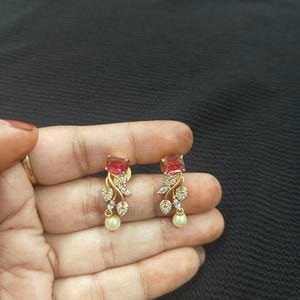 Op de afbeelding: Een paar goudkleurige oorbellen met een rechthoekige, roze edelsteen. De oorbellen hebben een bloemmotief met heldere kristallen en een bungelende parel. De oorbellen worden in een hand tegen een donkere achtergrond gehouden.