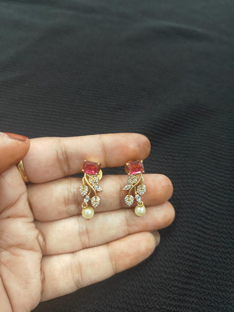 Op de afbeelding: Een paar goudkleurige oorbellen met een rechthoekige, roze edelsteen. De oorbellen hebben een bloemmotief met heldere kristallen en een bungelende parel. De oorbellen worden in een hand tegen een donkere achtergrond gehouden.