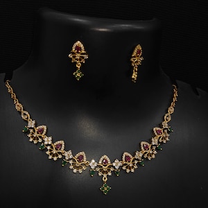 May include: A gold-toned necklace and earring set. The necklace features a series of floral-shaped pendants with red and white gemstones, interspersed with green gems. The earrings mirror the design, with a single red gem and green gem.
