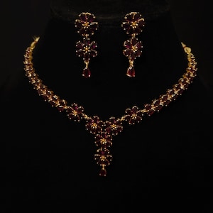 May include: A gold-toned necklace and earring set. The jewelry features a floral design with burgundy-colored gemstones. The necklace has a cascading floral pendant, and the earrings are a matching drop style. The set is displayed against a black background.