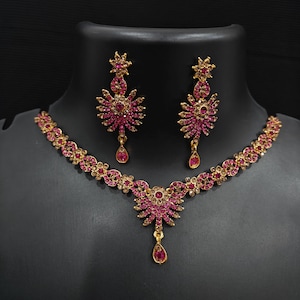 May include: A gold-coloured necklace and earring set with pink gemstones. The necklace features a floral design with a central pendant and matching teardrop earrings. The jewellery is displayed on a black mannequin.