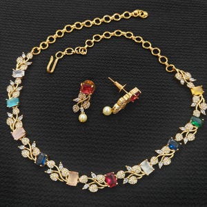 Op de afbeelding: Een goudkleurige ketting- en oorbellenset. De ketting heeft kleurrijke rechthoekige edelstenen en delicate blad- en kristalaccenten. De oorbellen hebben een druppelvormige edelsteen en een kleine parel. De sieraden staan tegen een zwarte achtergrond.