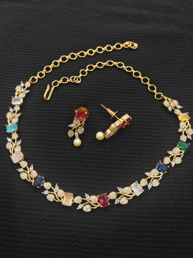 Op de afbeelding: Een goudkleurige ketting- en oorbellenset. De ketting heeft kleurrijke rechthoekige edelstenen en delicate blad- en kristalaccenten. De oorbellen hebben een druppelvormige edelsteen en een kleine parel. De sieraden staan tegen een zwarte achtergrond.