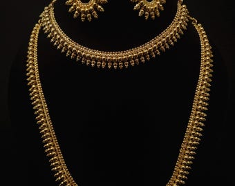 May include: A gold-colored jewelry set featuring a necklace and matching earrings. The necklace has two strands, one shorter and one longer, both with a beaded design. The earrings have a sunburst design. The set is displayed against a black background.