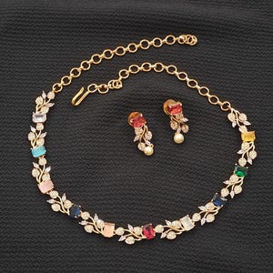 Op de afbeelding: Een goudkleurige ketting- en oorbellenset. De ketting heeft een reeks kleurrijke rechthoekige edelstenen, waaronder blauw, roze en rood, afgewisseld met delicate blad- en kristalaccenten. De bijpassende oorbellen hebben een vergelijkbaar ontwerp met een pareldruppel.