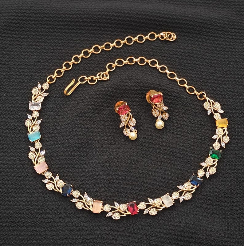 Op de afbeelding: Een goudkleurige ketting- en oorbellenset. De ketting heeft een reeks kleurrijke rechthoekige edelstenen, waaronder blauw, roze en rood, afgewisseld met delicate blad- en kristalaccenten. De bijpassende oorbellen hebben een vergelijkbaar ontwerp met een pareldruppel.