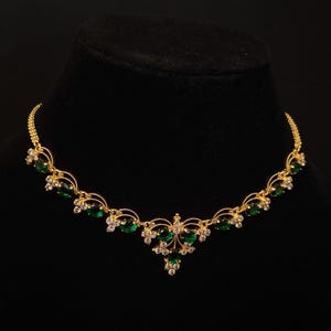 May include: A gold necklace with emerald green and clear gemstone detailing. The necklace has a delicate design with a central pendant and symmetrical stone arrangement. The necklace is displayed on a black mannequin.