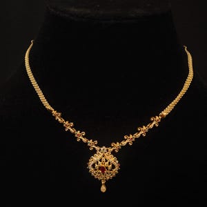 Puede incluir: Un collar dorado con un colgante central con una piedra roja rodeada de pequeñas piedras blancas. La cadena tiene un diseño intrincado con elementos florales. El collar se exhibe en un maniquí negro, mostrando su elegancia.