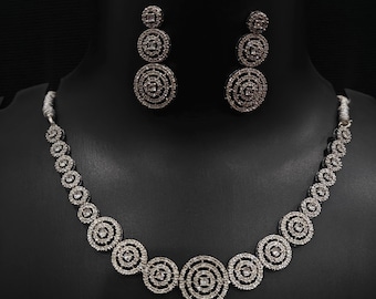 May include: A silver-toned jewelry set featuring a necklace and matching earrings. The necklace and earrings have a circular design with concentric circles and sparkling accents. The set is displayed on a black mannequin.