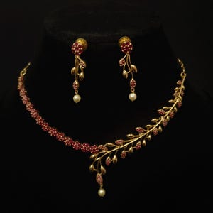 Peut inclure: Un ensemble de bijoux doré comprenant un collier et des boucles d'oreilles assorties. Le collier et les boucles d'oreilles présentent un motif floral avec des pierres rouges et de petites perles blanches. L'ensemble est présenté sur fond noir.