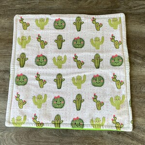 May include: A square, white baby blanket with a repeating pattern of green cacti with pink flowers. The blanket has a lime green border and a white border. The cacti have smiling faces.