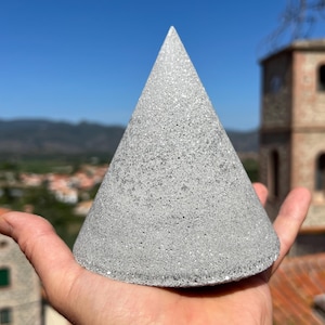 Peut inclure: Objet conique gris avec une surface texturée, tenu dans une main devant un ciel bleu et un bâtiment. Le cône a un sommet pointu et une base large, avec un aspect moucheté.
